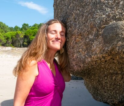 Carmen en la playa con un vestido rosa tocando una roca enorme en la que apoya su cara