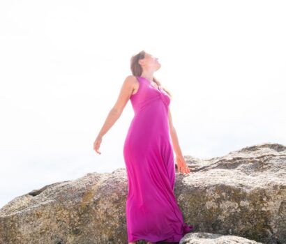 Carmen con un vestido rosa subida a una roca en la playa mirando al sol.