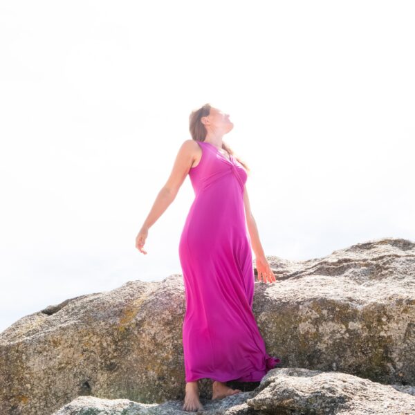 Carmen con un vestido rosa subida a una roca en la playa mirando al sol.